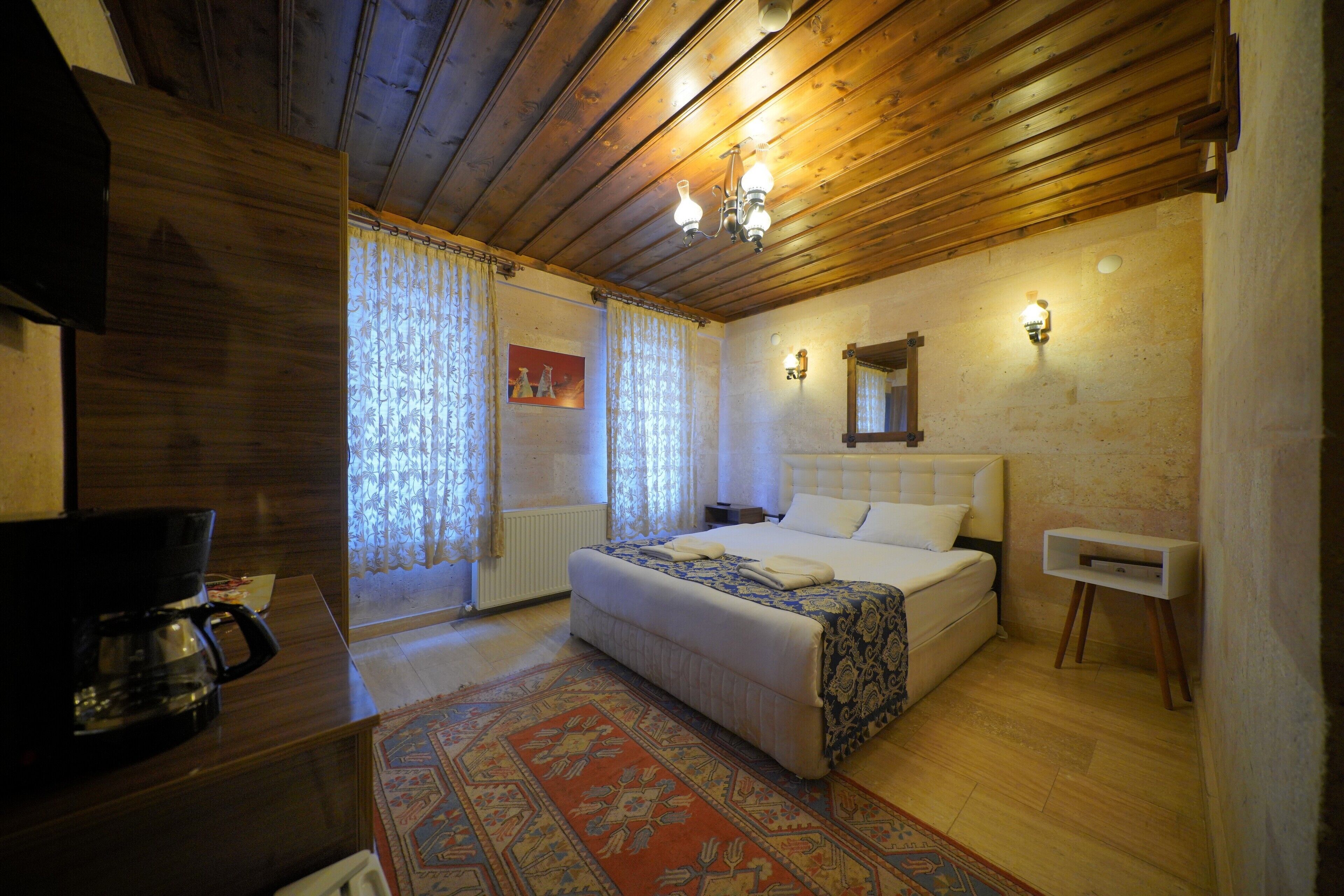 A bedroom with a wooden ceiling, a bed with a white headboard, a bedside table, a nightstand, a wall-mounted lamp, and a patterned rug.
