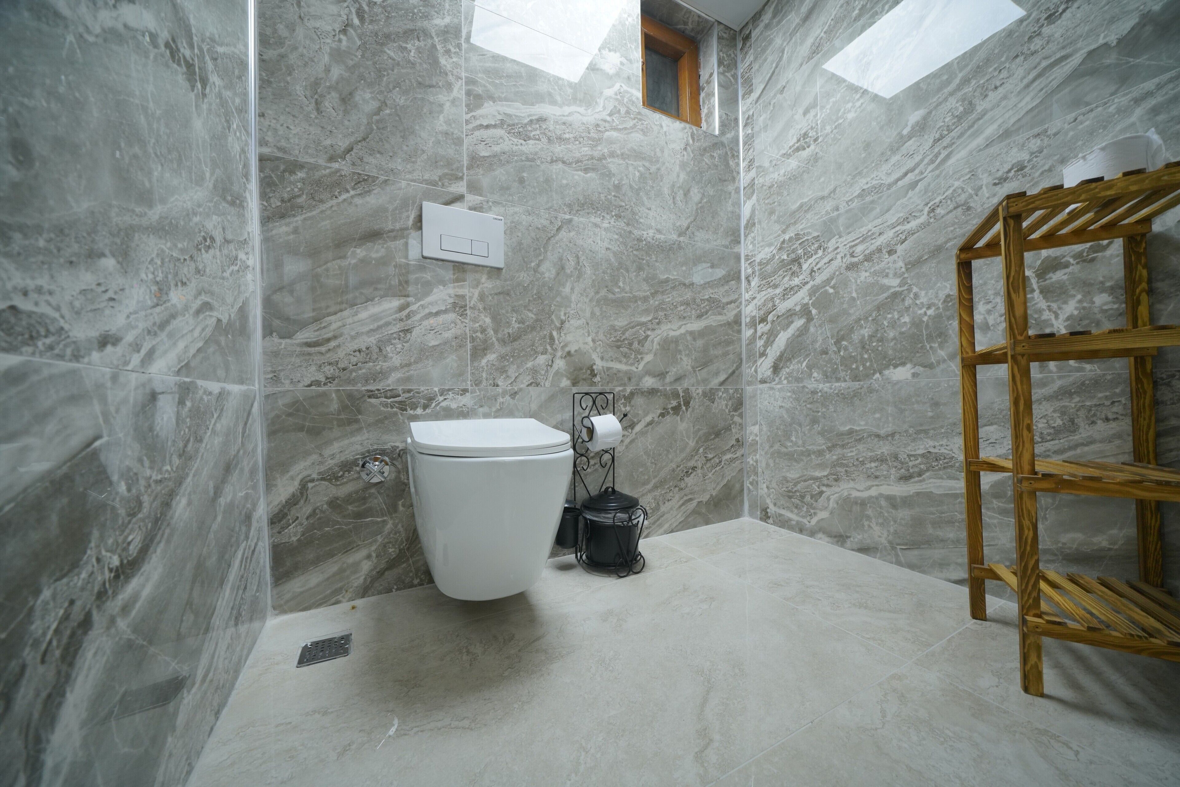 A bathroom with a marble wall, a white toilet, a wooden shelf, and a skylight.