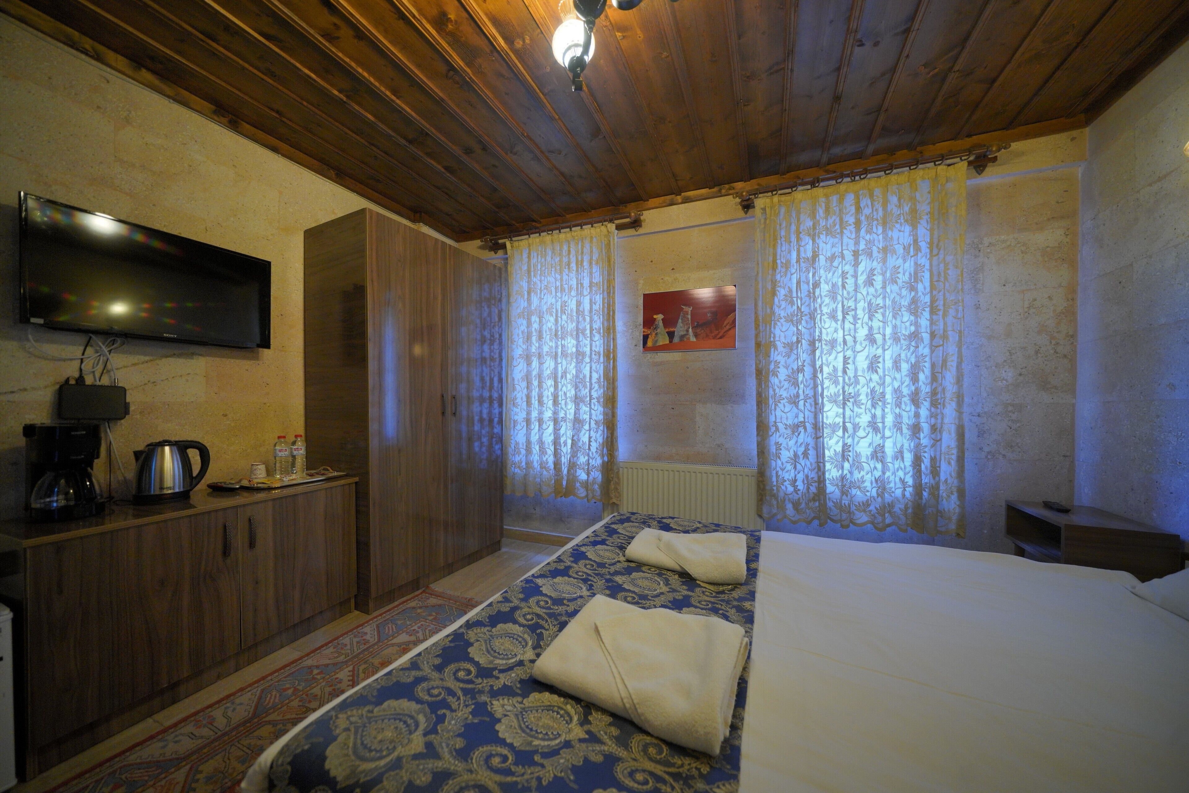 A bedroom with a wooden ceiling, a TV mounted on the wall, a bed with a patterned bedspread, a wardrobe, and a radiator.