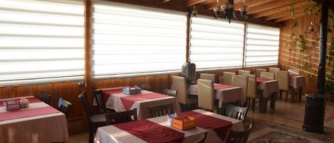 A restaurant with wooden beams, tables with red and white tablecloths, and a large window with blinds.