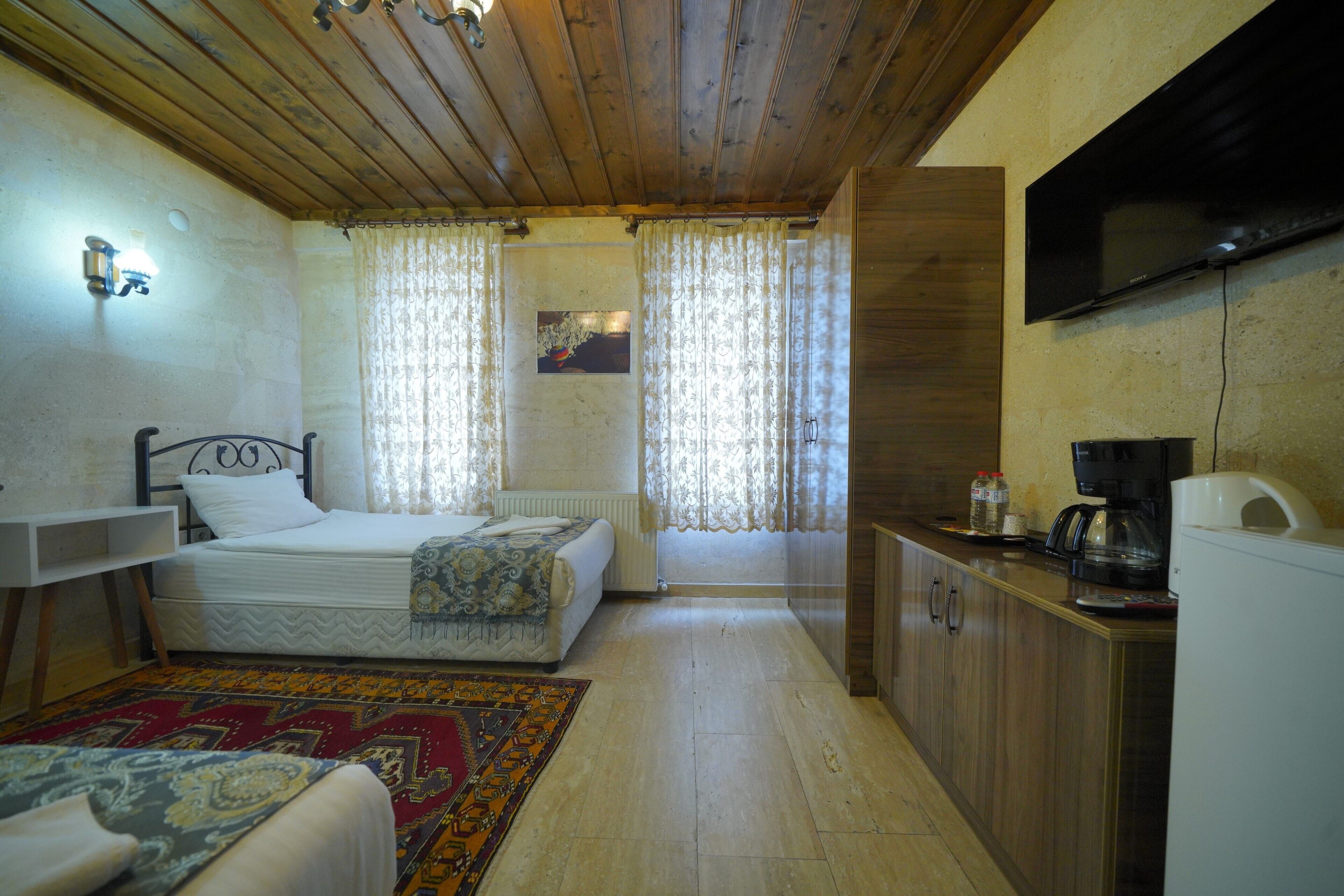 A bedroom with a bed, a desk, a television, and a kitchen area with a refrigerator and coffee maker.