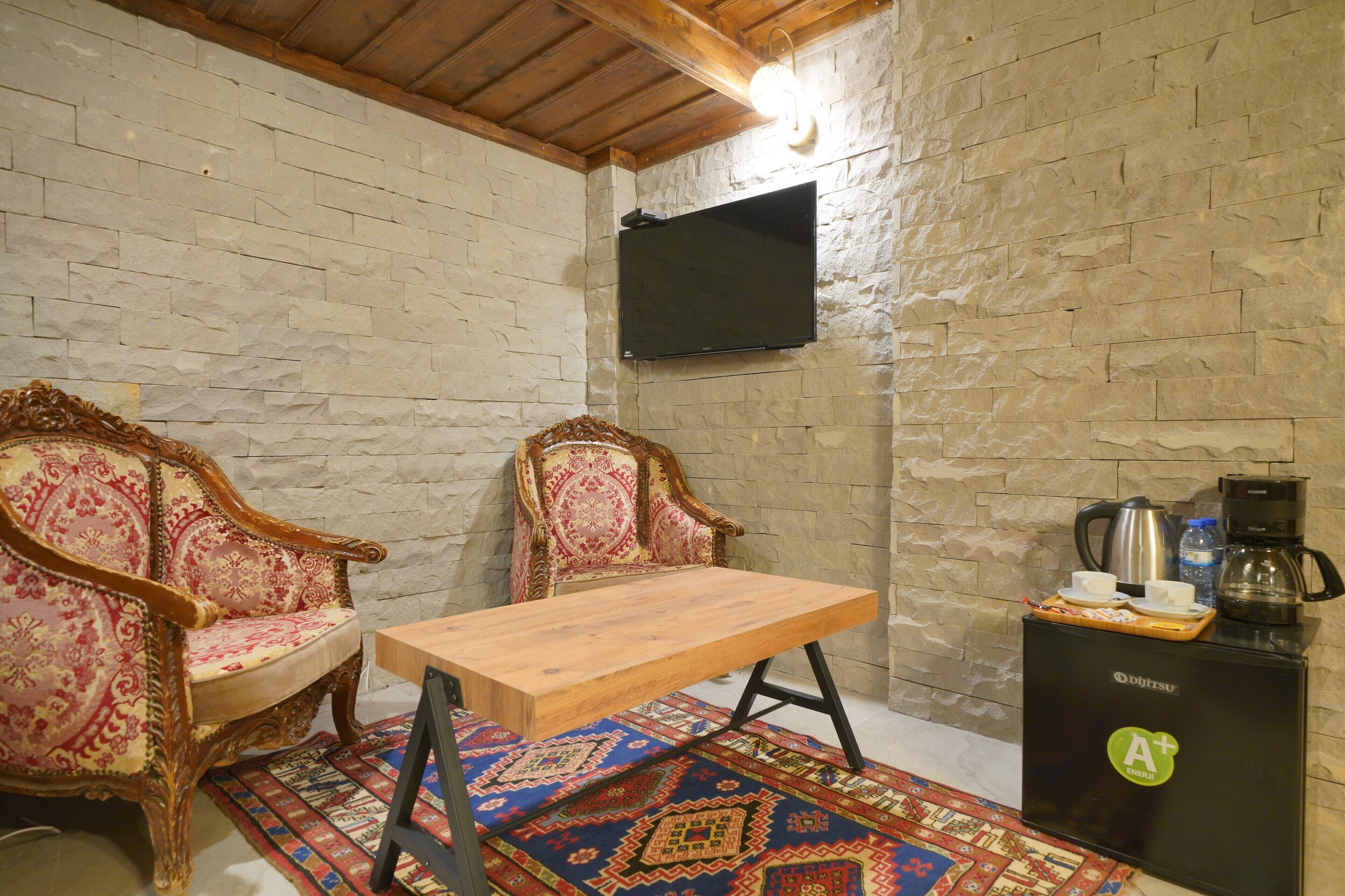 Une pièce comprenant deux fauteuils richement décorés, une table en bois, un téléviseur fixé à un mur en pierre, ainsi qu’un coin café avec une bouilloire et des tasses.