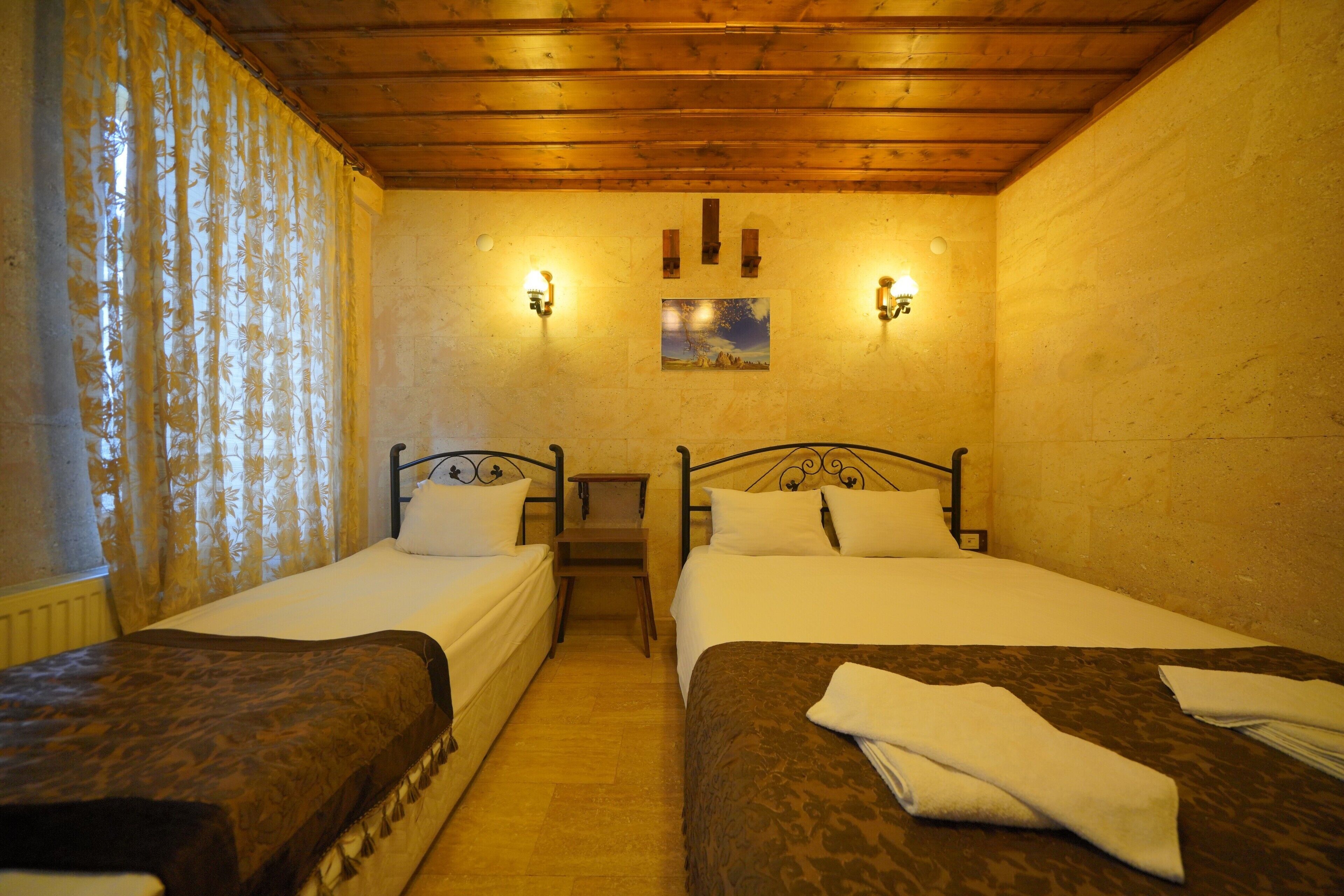 A room with two beds, wooden ceiling beams, wall-mounted lights, and a small painting on the wall.