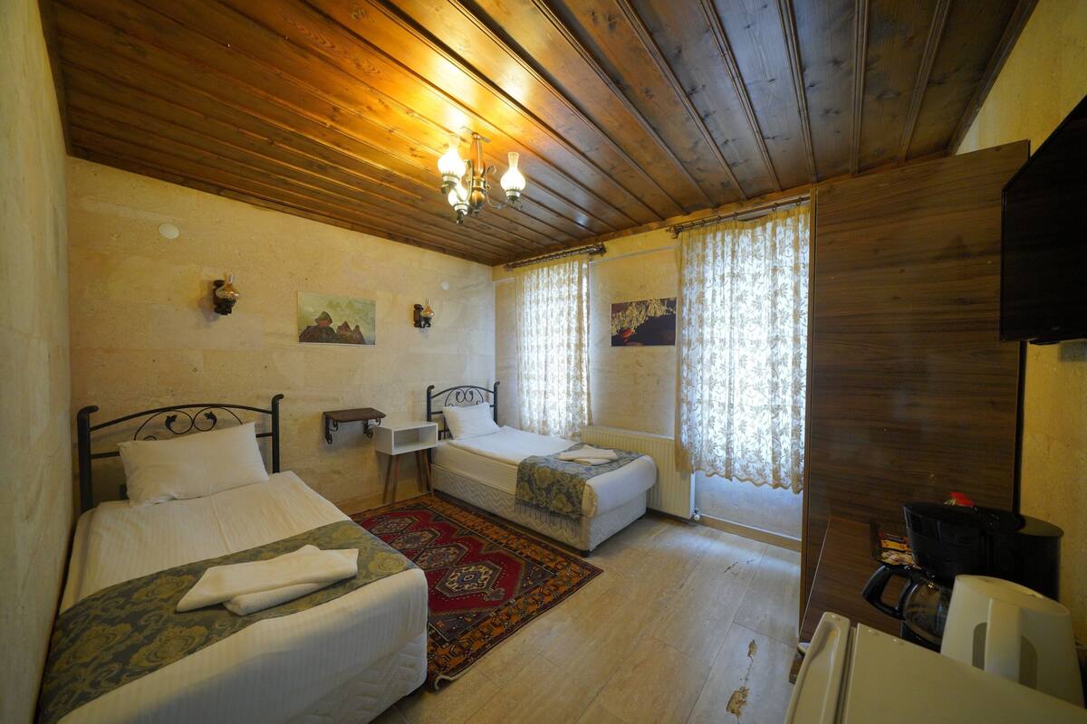 A room with two beds, a wooden ceiling, a chandelier, a radiator, and a window with curtains.