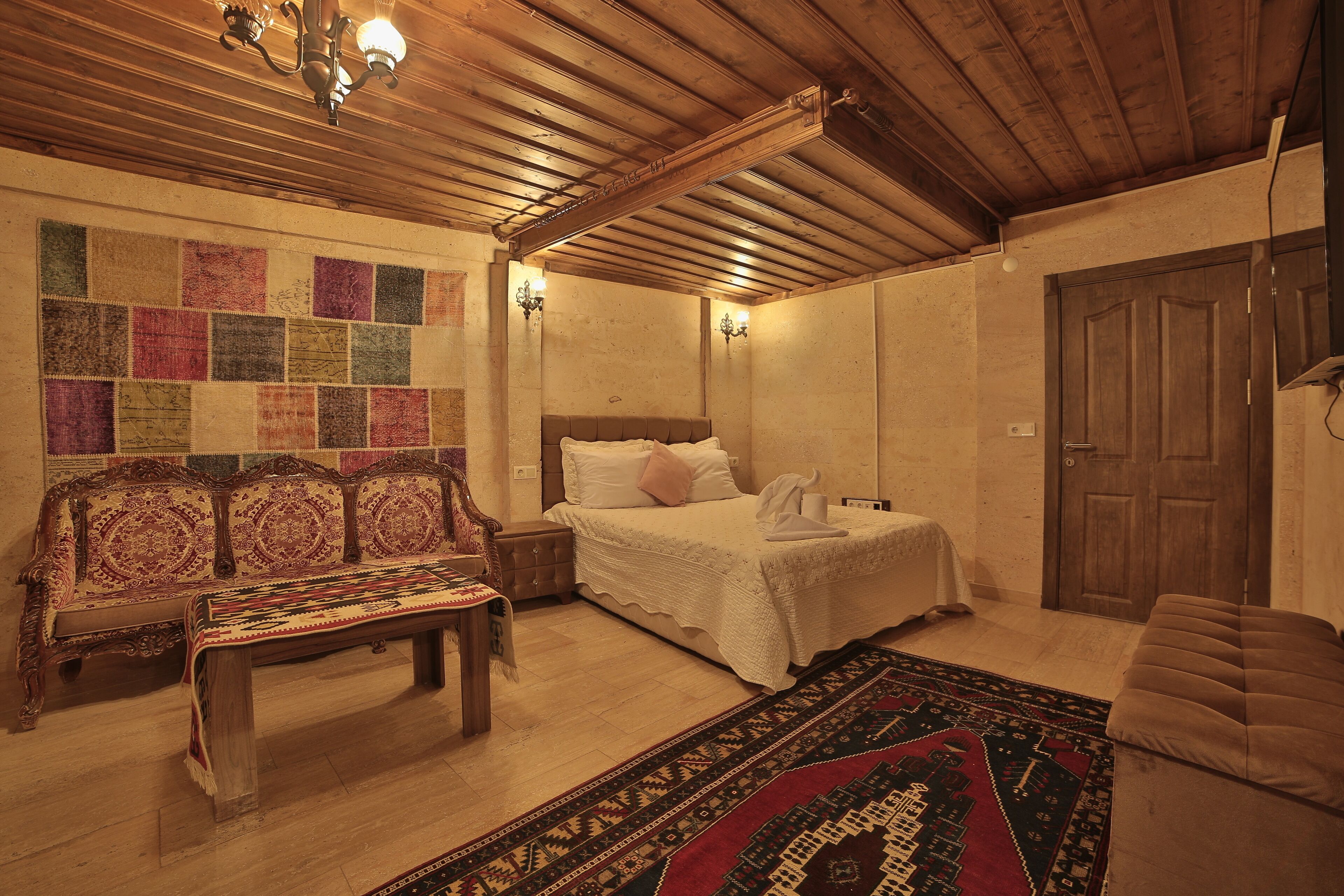 A cozy room with a wooden ceiling, a bed with white linens, a bench, and a colorful rug.