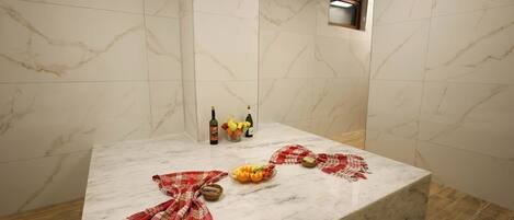A marble-tiled bathroom with a sink, bottles, and a bowl of fruit.