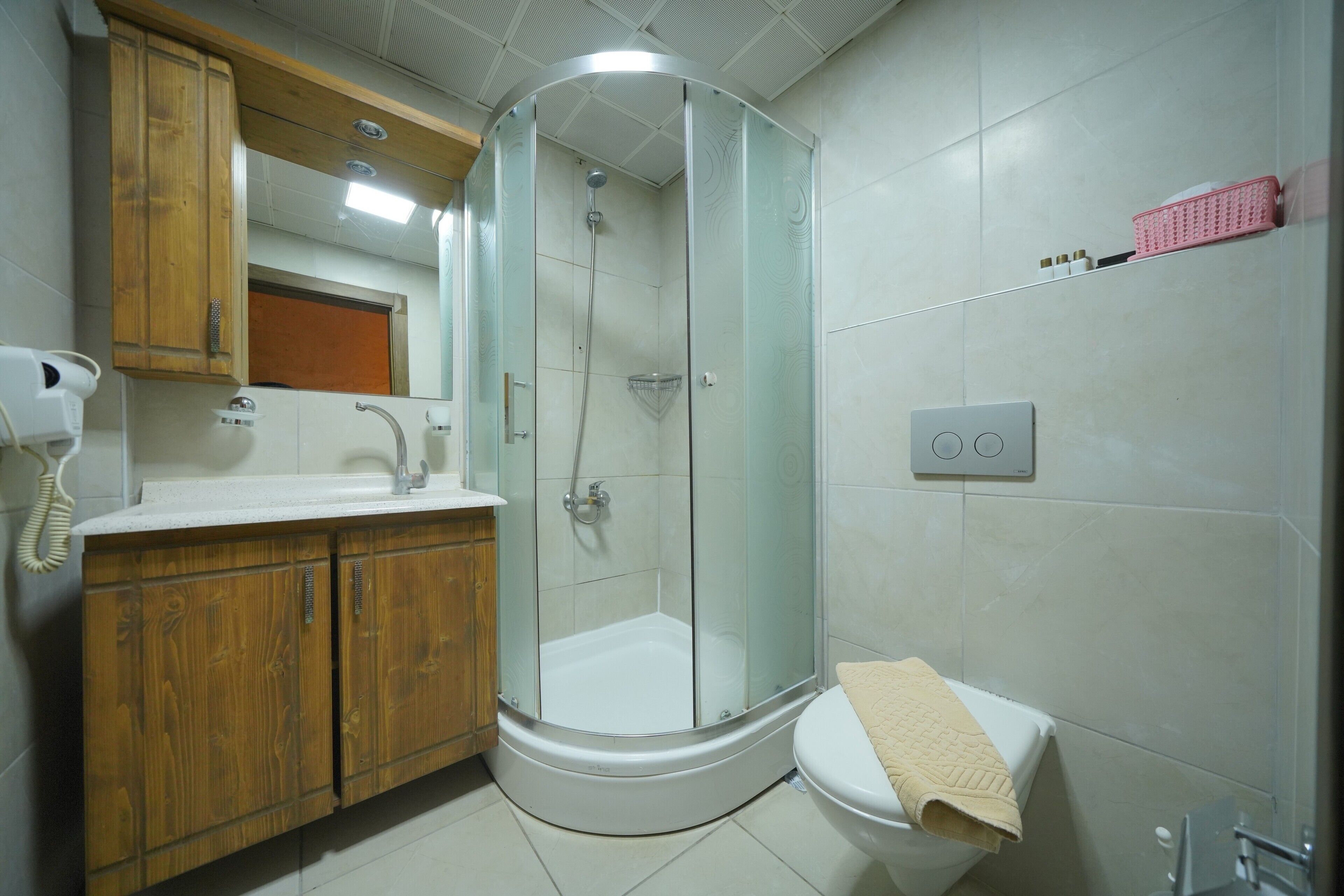 Un baño moderno con ducha de vidrio, tocador de madera, inodoro y una cesta rosa en la pared.