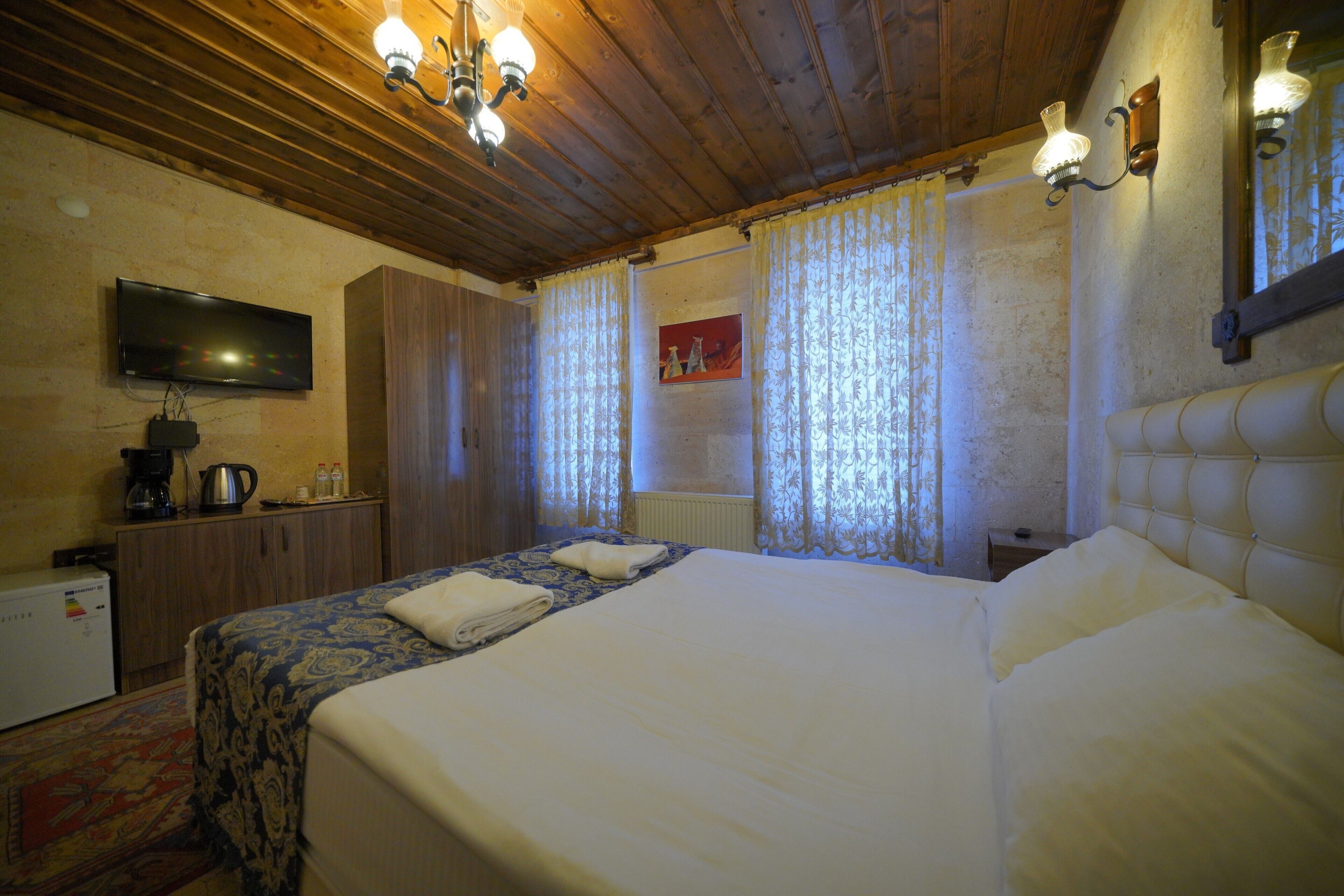 Habitación de hotel con techo de madera, televisor montado en la pared, cama con cabecera, mesita de noche y ventana con cortinas.
