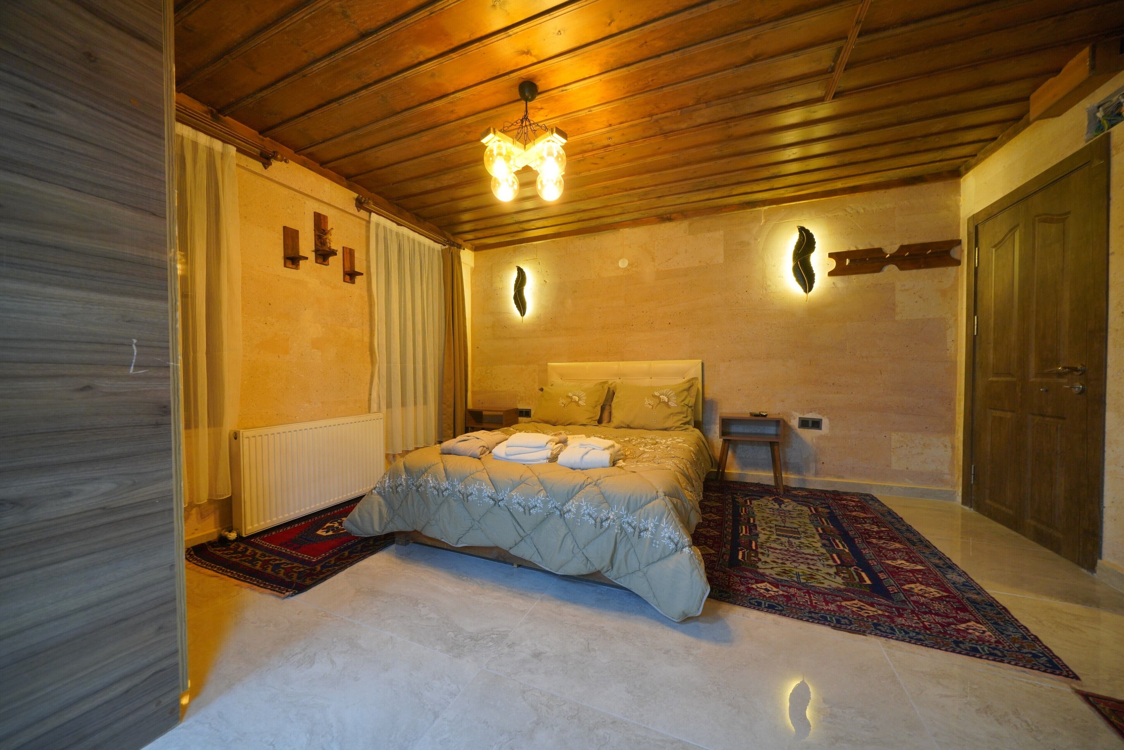 A bedroom with a wooden ceiling, a bed with a beige duvet, a nightstand, a wall-mounted light, and a door.