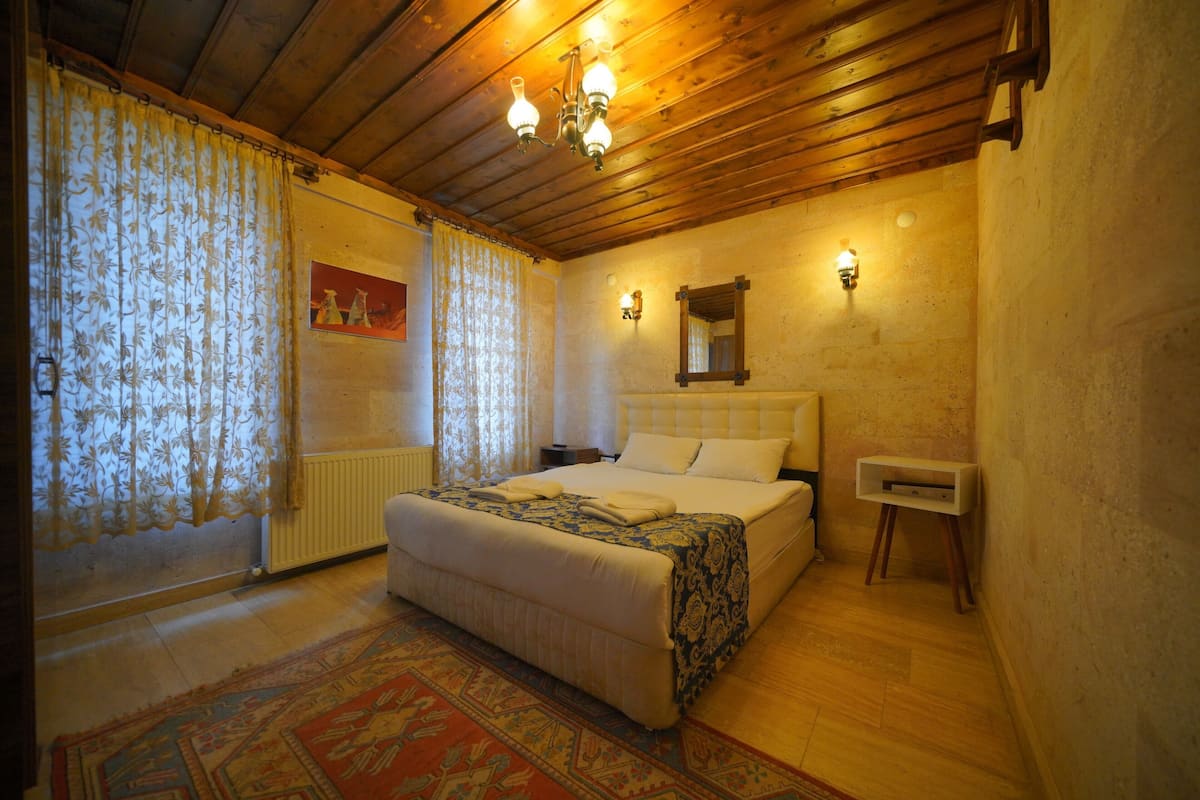A bedroom with a wooden ceiling, a bed with a patterned bedspread, a nightstand, a wall-mounted mirror, and a small bedside table.