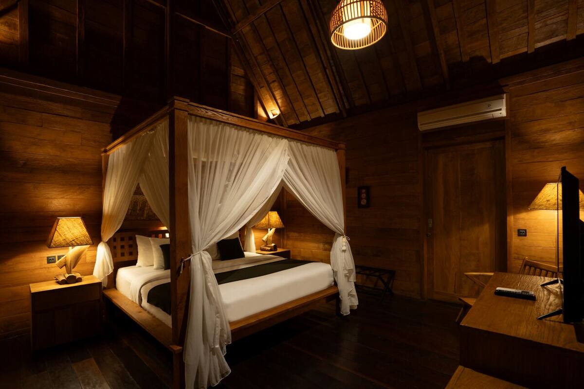 A four-poster canopy bed with white linens, a wooden nightstand with a lamp, and a wooden table with a remote control.