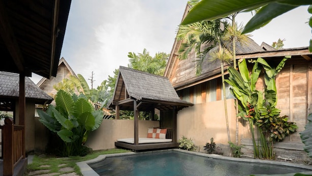Un área de piscina con cubierta de madera, un rincón de descanso con cojines y plantas tropicales.