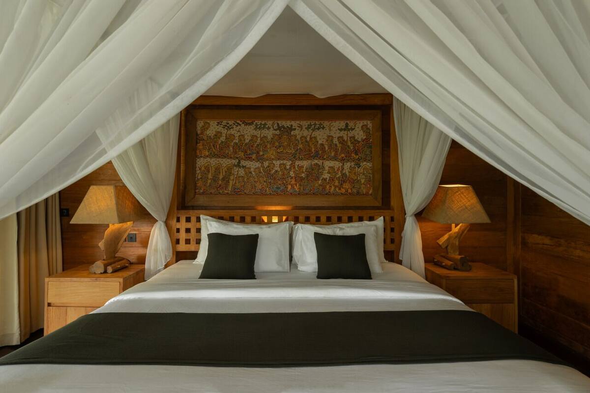 A bed with white linens, two dark pillows, and a green throw blanket. There are two bedside tables with lamps. The room has a wooden headboard and a large tapestry above the bed.