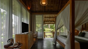 Habitación con piso de madera, cama con cortinas blancas, televisor sobre un mueble de madera y vista a un jardín por la ventana.