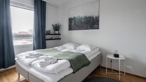 Un letto rifatto con lenzuola bianche e una coperta verde, un comodino con una lampada, una mensola con piante e un quadro raffigurante una foresta al muro.