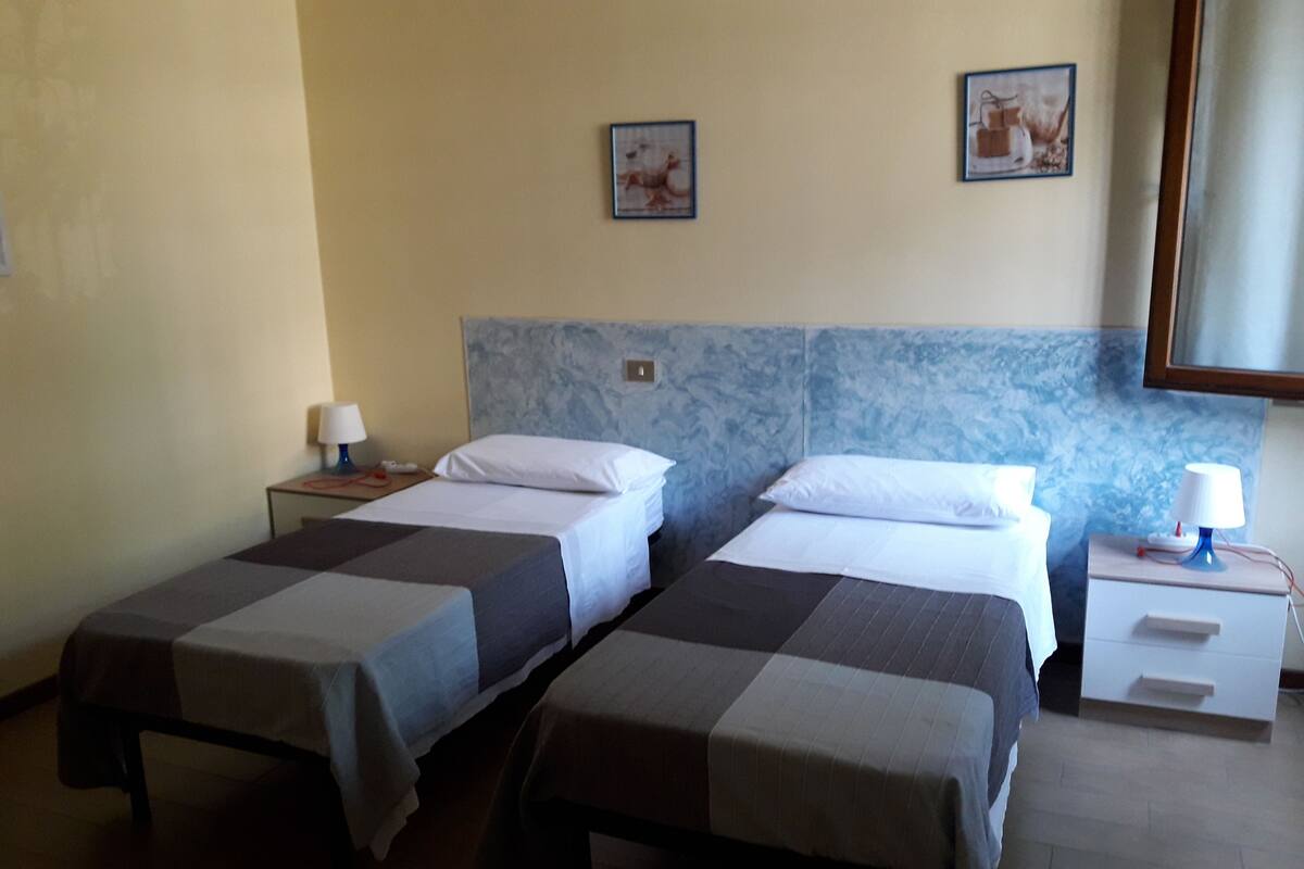 A hotel room with two single beds, a nightstand with a lamp, and a wall-mounted picture.