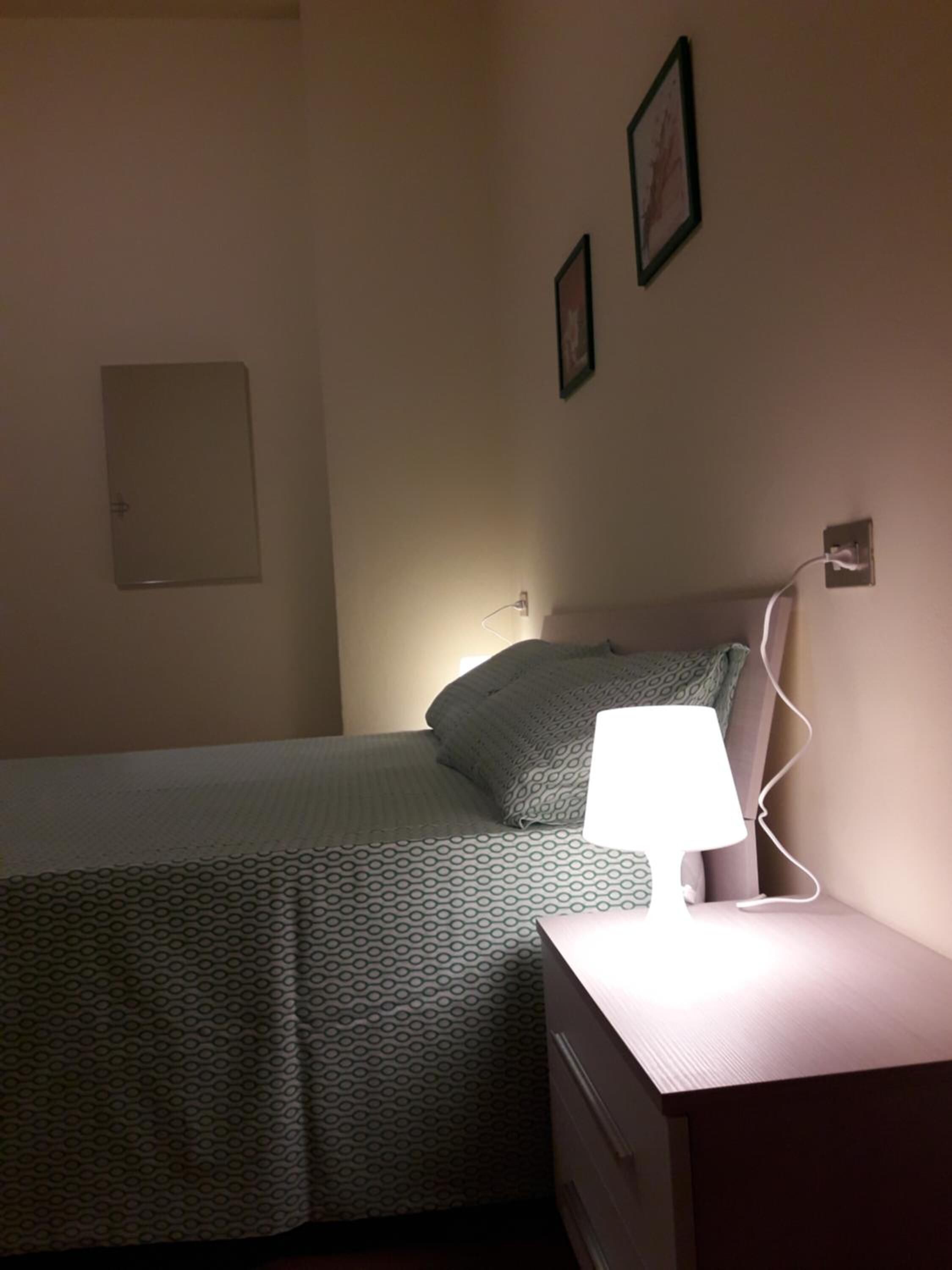 A bedroom with a bed, a bedside table with a lamp, a power outlet, and two framed pictures on the wall.