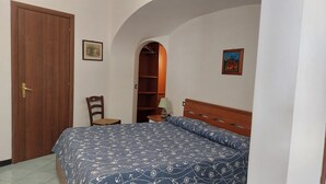 Un dormitorio con puerta de madera, una silla, una cama con colcha estampada, una mesita de noche, una lámpara y un armario.