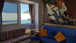 Una habitación con un ventanal grande que da a una playa, un sofá azul con cojines, un banco a rayas y un tapiz decorativo.