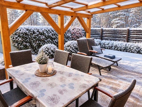 Ein überdachter Außenbereich mit Tisch und Stühlen, umgeben von schneebedeckter Bepflanzung und einem Holzzaun.