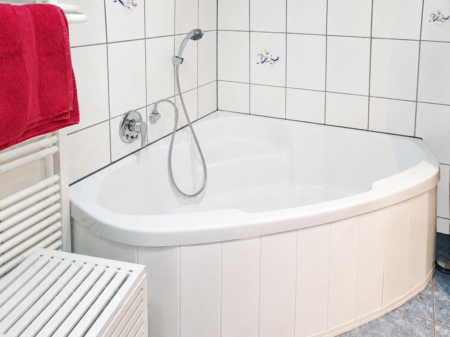A white bathtub with a shower head and control knobs, a red towel on a white radiator, and white tiled walls with decorative tiles.