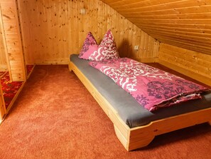 Yksinkertainen puinen sänky, jossa on harmaa patja ja vaaleanpunaisin kuvioin varustettu vuodevaatesetti. Huoneessa on puuseinät ja kalteva katto.