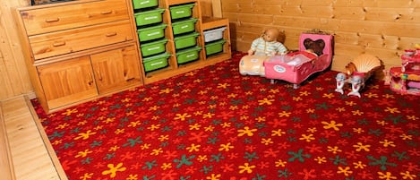 Una habitación con televisión sobre un mueble de madera, contenedores verdes para guardar objetos, una alfombra roja estampada y juguetes infantiles.