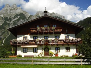 A traditional chalet with a wooden balcony, a staircase leading to the entrance, and a mountain backdrop.