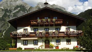 A traditional chalet with a wooden balcony, a staircase leading to the entrance, and a mountain backdrop.