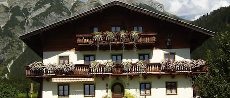 A traditional chalet with a wooden balcony, a staircase leading to the entrance, and a mountain backdrop.