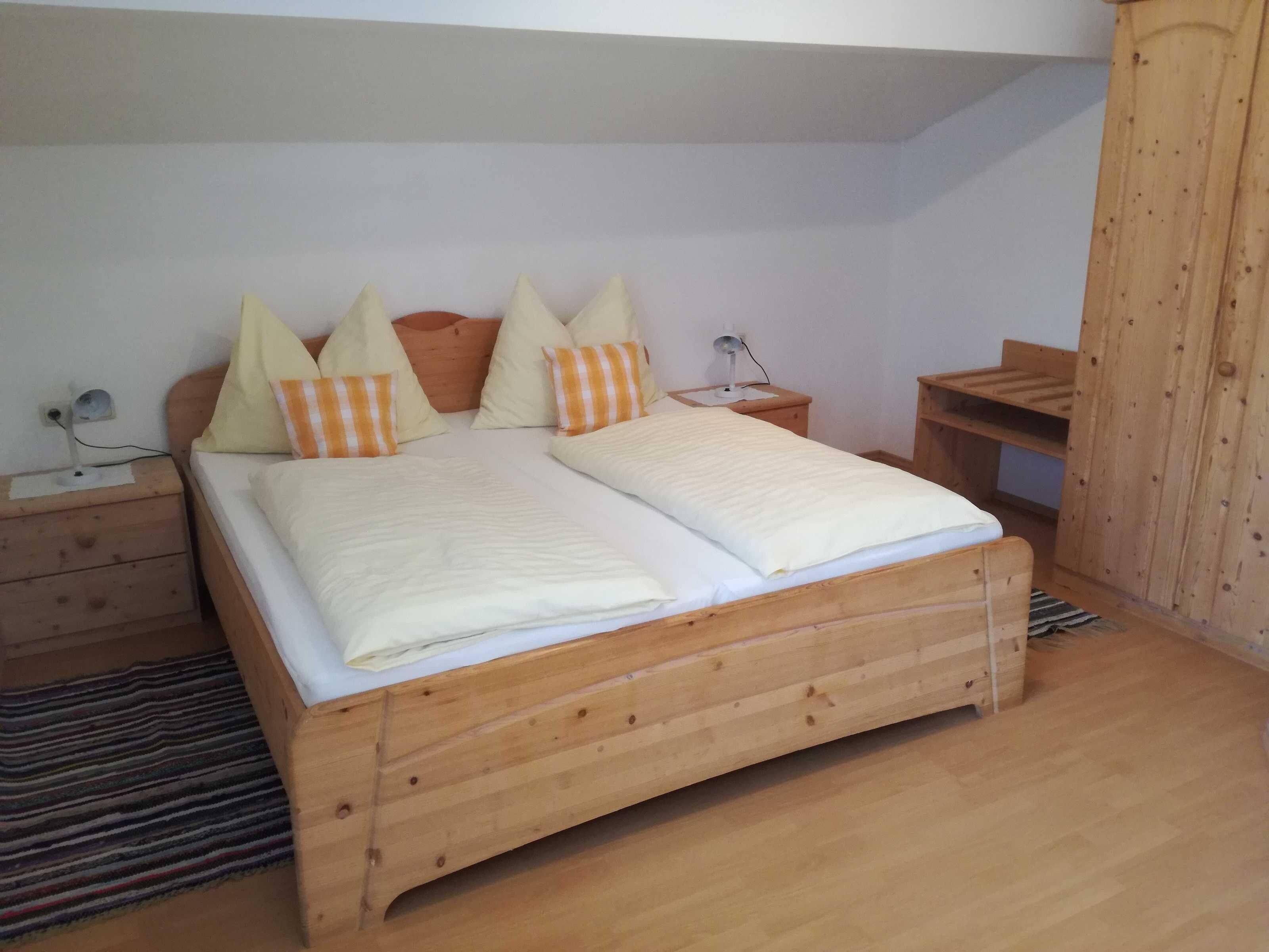 A wooden bed with a white mattress and striped pillows, a bedside table with a lamp, and a wooden wardrobe.