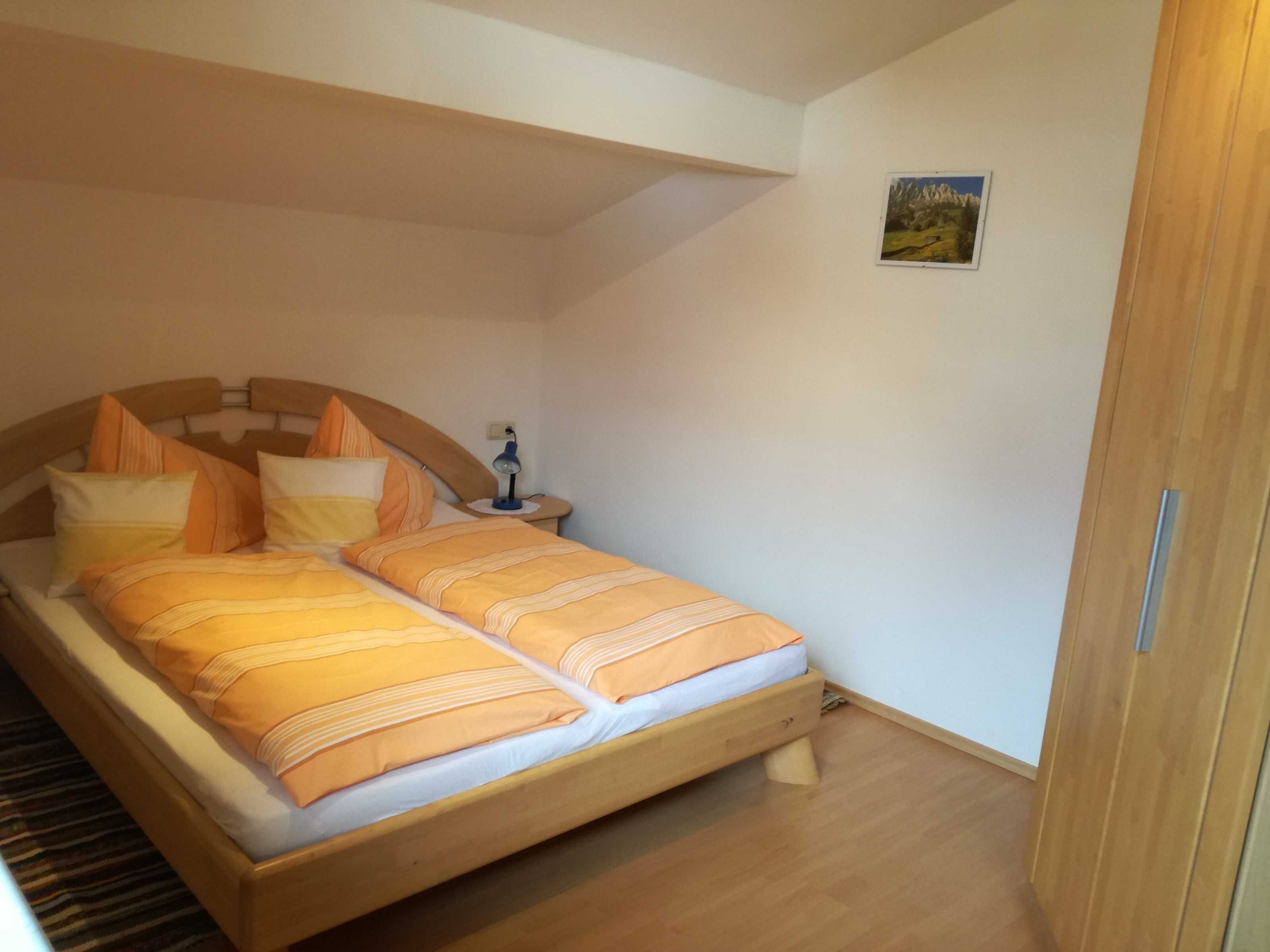 A single bed with a wooden frame and orange bedding, a bedside table with a lamp, and a small painting on the wall.