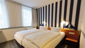 A hotel room with a large bed, two bedside tables with lamps, a window with curtains, and a striped wall. - Hotel DH VEC (Vechta)