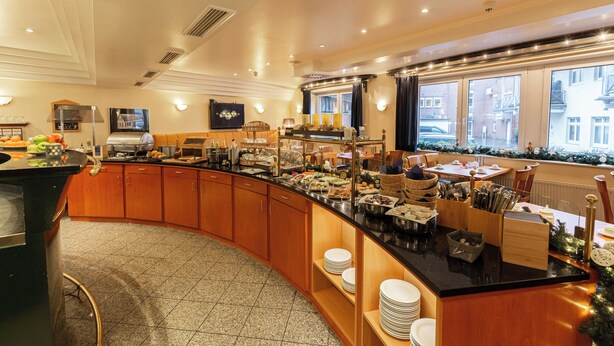 A breakfast bar with various food items and utensils, a seating area with tables and chairs, and a view of the city through large windows. - Hotel DH VEC (Vechta)