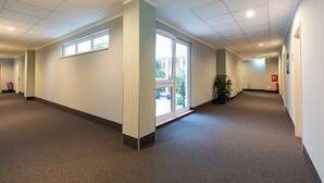 A spacious hallway with carpeted floors, white walls, and ceiling lights. There are windows along the walls and a door leading outside. - Hotel DH VEC (Vechta)