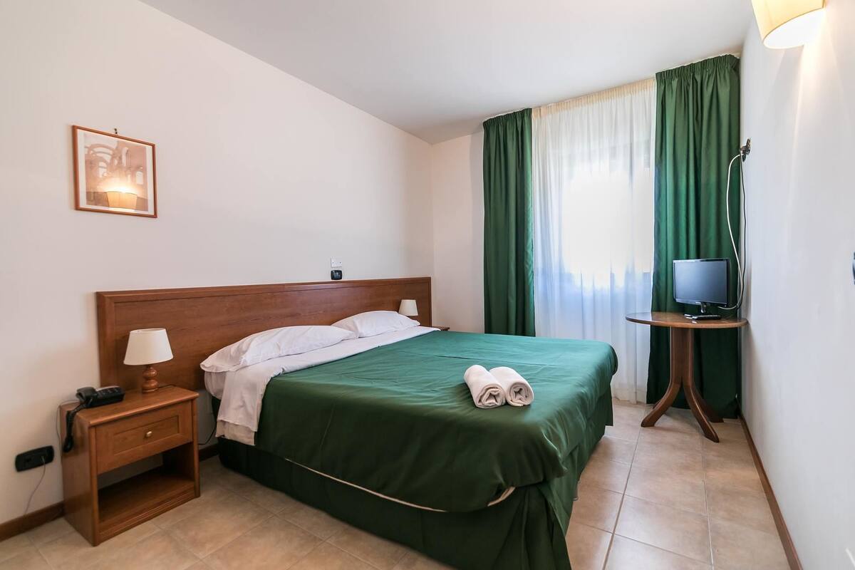A hotel room with a bed, a bedside table with a lamp, a TV on a small table, and a window with green curtains.