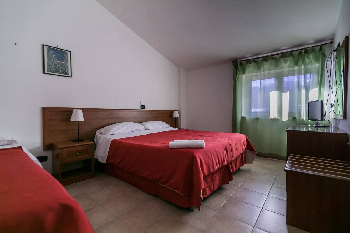A hotel room with two beds, a desk with a computer, and a window with green curtains.