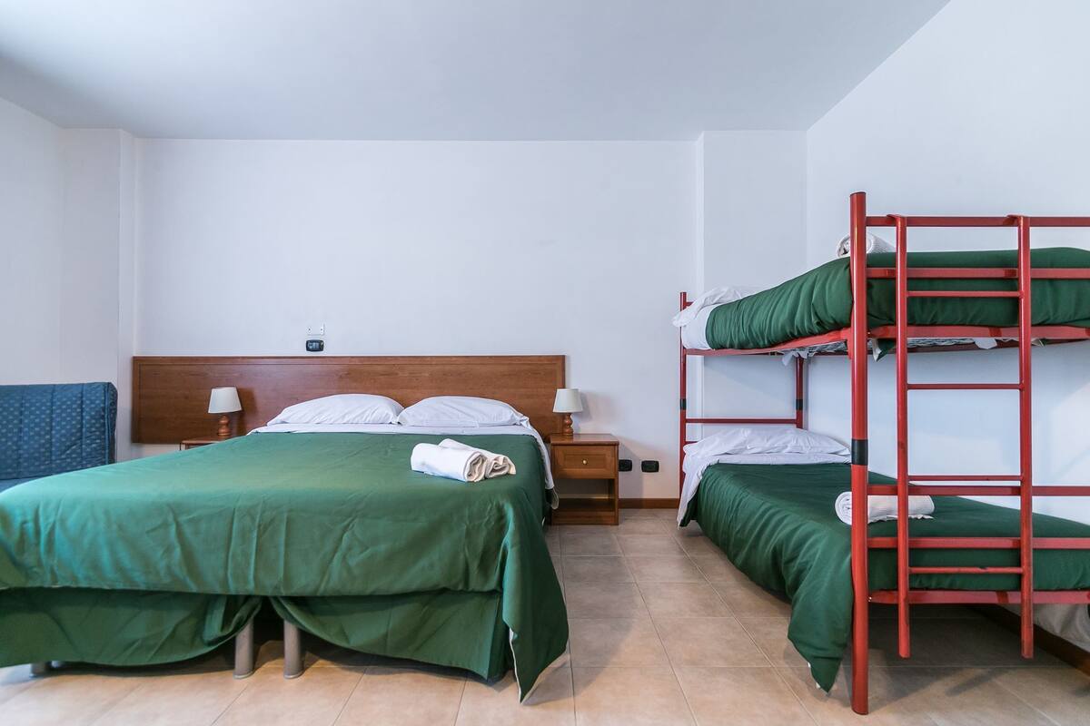 A dormitory room with bunk beds, a single bed, wooden headboard, bedside tables with lamps, and a blue patterned wall.
