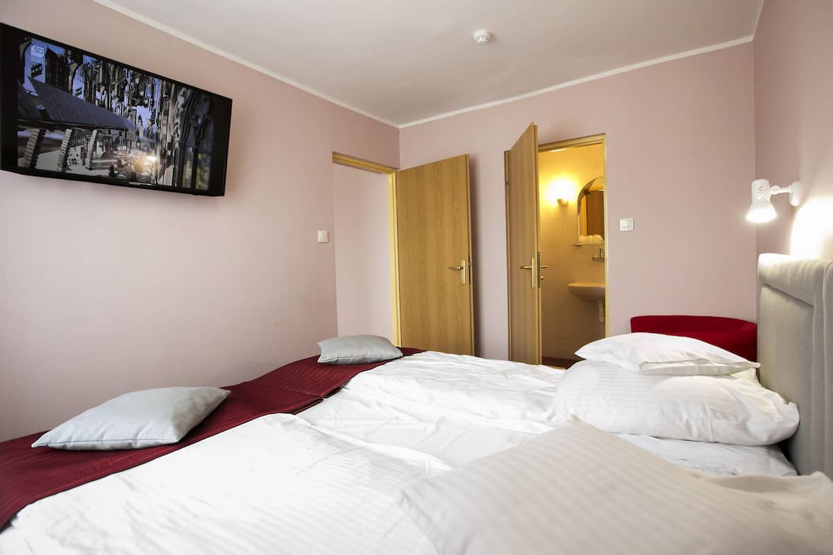 Quarto com cama, almofadas, uma cadeira vermelha, um guarda-roupa de madeira e uma televisão presa à parede.
