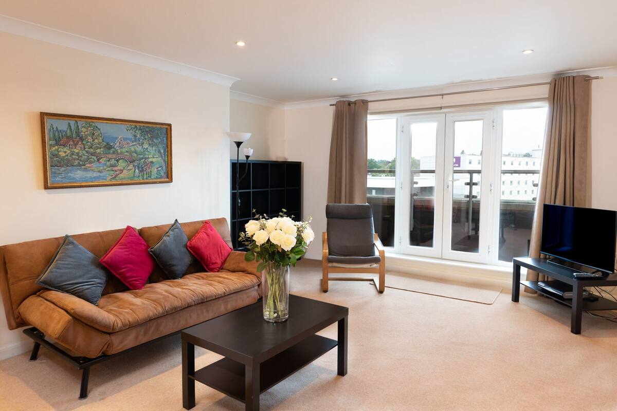 A living room with a brown leather sofa, a black coffee table, a vase with white flowers, a black TV stand, and a painting on the wall.