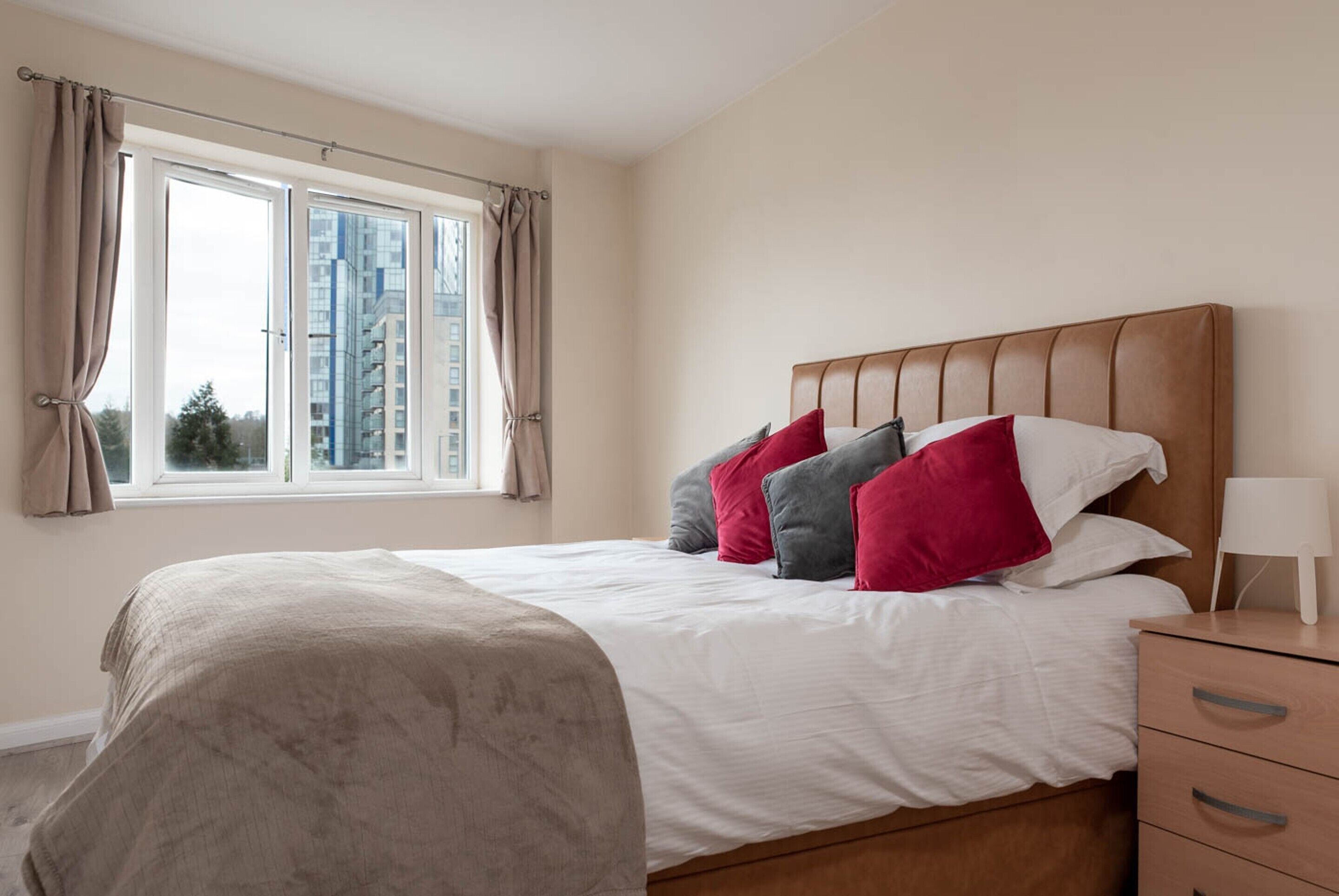 A bedroom with a large bed, a wooden nightstand, a window with curtains, and a view of buildings outside.