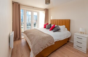 A modern bedroom with a large bed, a bedside table, and a view of the city through a large window. - Abodebed - Handleys Court (Hemel Hempstead)