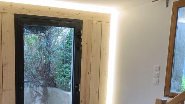A room with a wooden bench, a door leading outside, and a window with a view of trees.