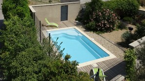 A pool with a wooden deck, surrounded by lush greenery and a small building with a tiled roof.
