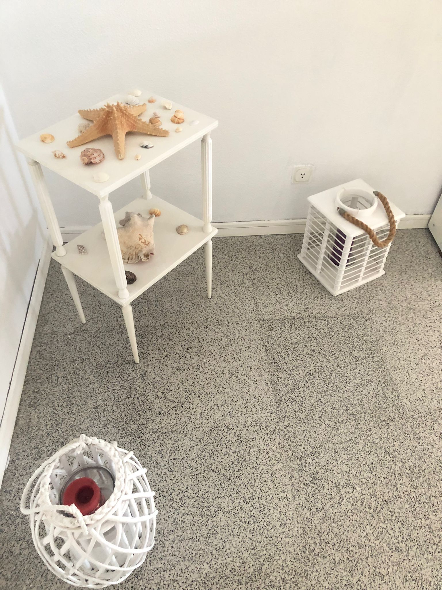 Een witte drielaagse plank met schelpen en een zeester, een witte mand met touwkoord, en een wit geweven mandje op een gevlekt tapijt.