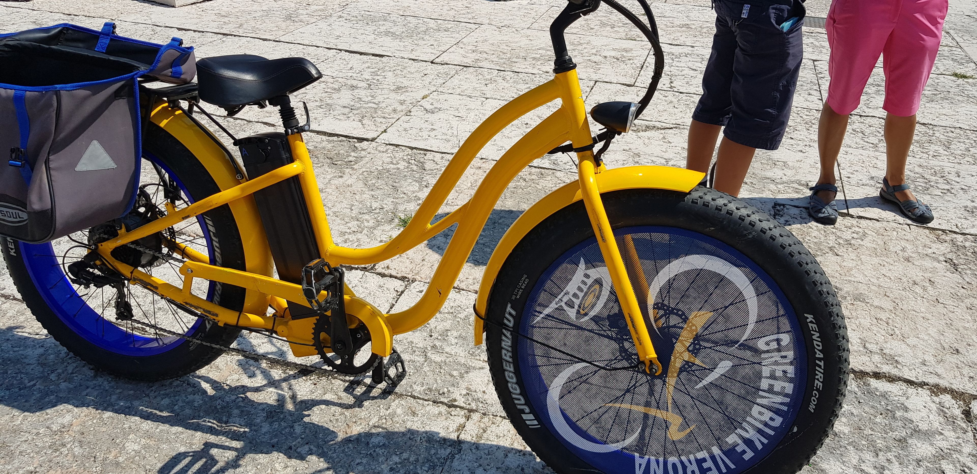 Una bicicleta amarilla con un bolso de almacenamiento y una cesta delantera, estacionada sobre una superficie pavimentada.