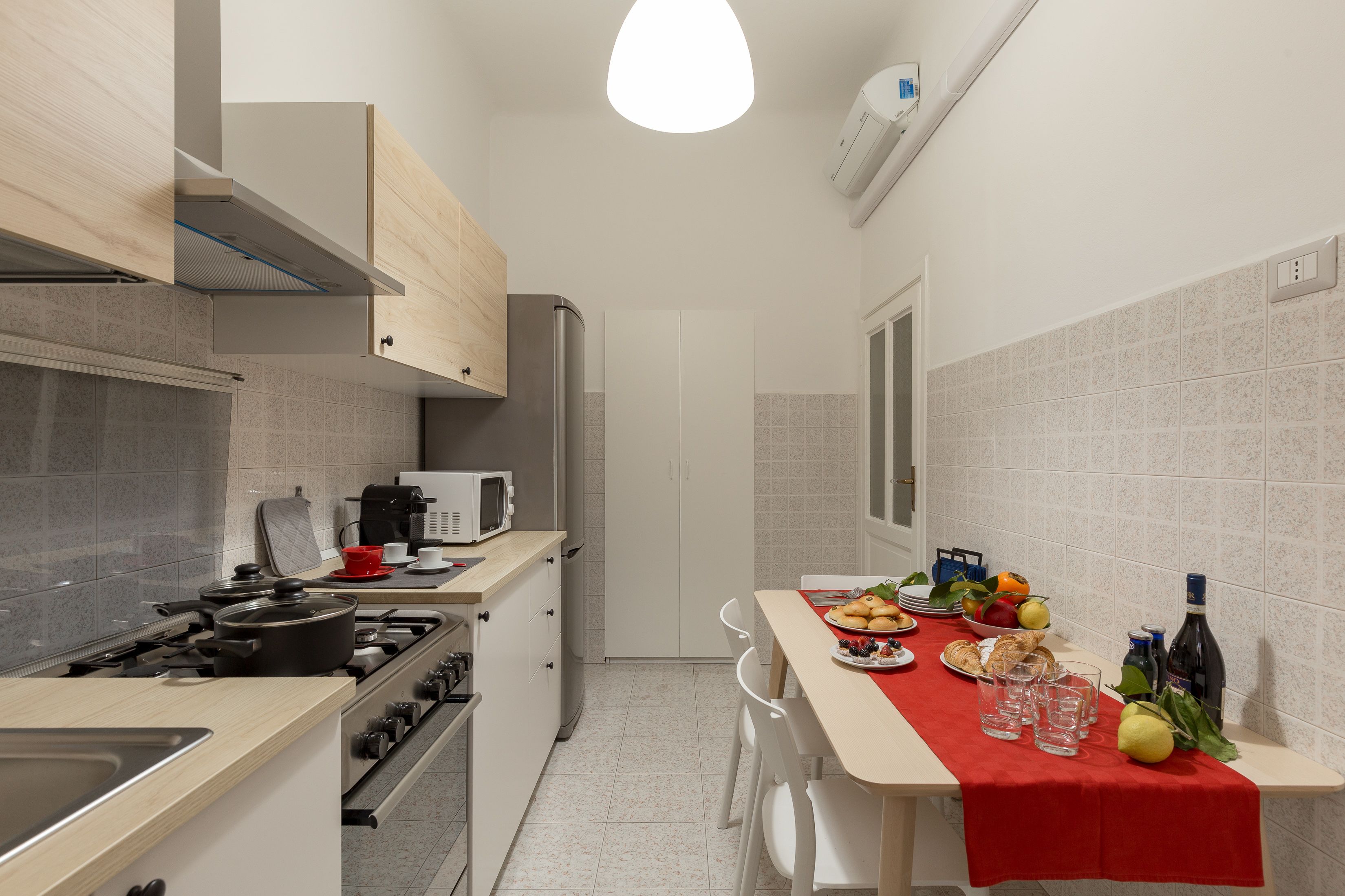 Una cocina compacta con estufa, microondas y armarios. Hay una mesa de comedor con mantel rojo y diversos alimentos.