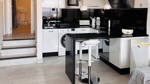 Een moderne keuken met een zwart aanrecht, witte kastjes, een magnetron en een spoelbak. Twee hanglampen en een stoel in de deuropening.