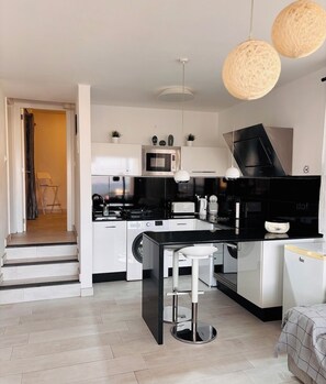 A modern kitchen with a black countertop, white cabinets, a microwave, and a sink. There are two pendant lights and a chair in the doorway.