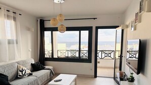 Een moderne woonkamer met een bank, een salontafel en een televisie. Grote ramen bieden uitzicht op zee en gebouwen.